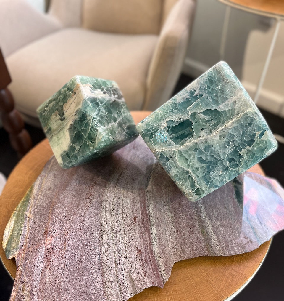 Two green and purple Fluorite cube stones on a wooden surface with a blurred background.