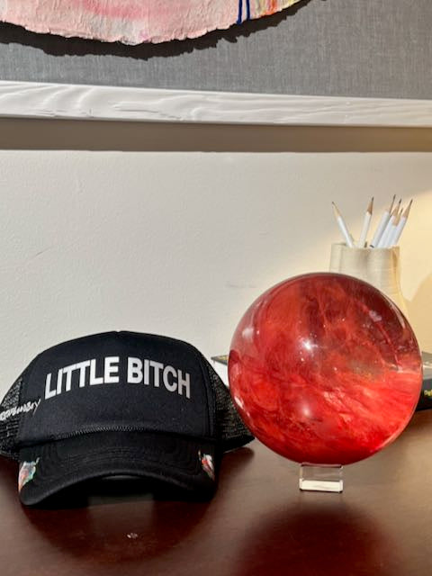 Cherry quartz sphere on desk next to baseball cap with words 
Little Bitch on it