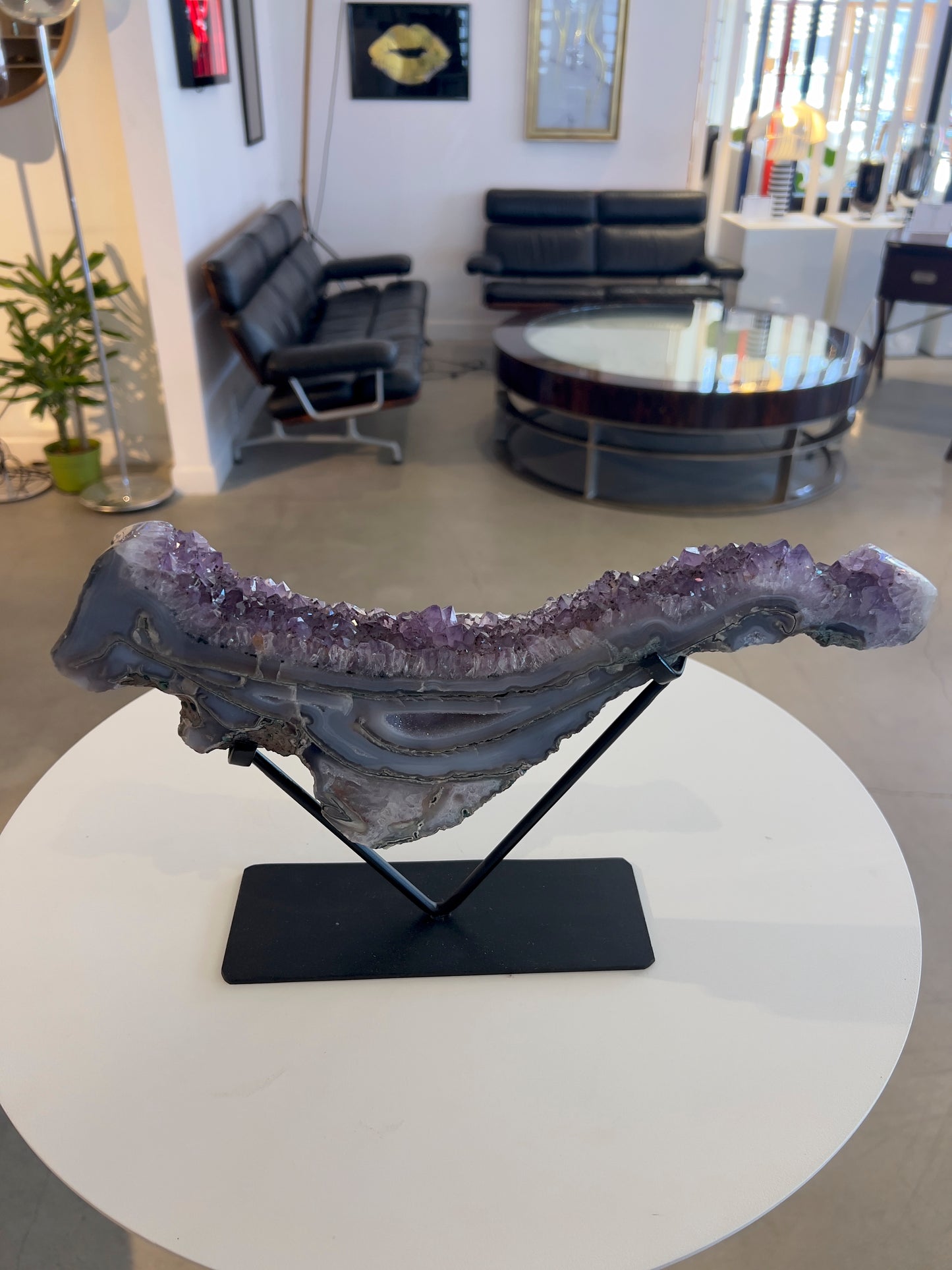 An Amethyst Agate Druzy wave on a black stand, displaying layered patterns of purple and blue with a crystalline structure. shown on white table with living room scene in distance