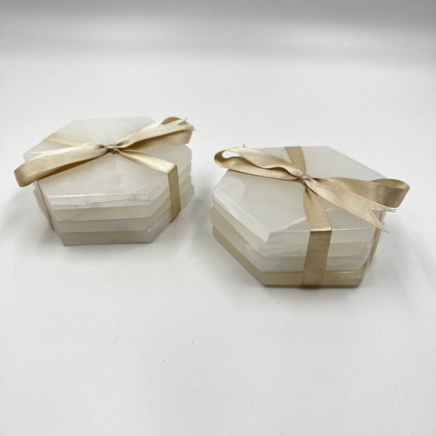 Two clear hexagonal coasters with white aragonite material and gold ribbon bows on a white background.