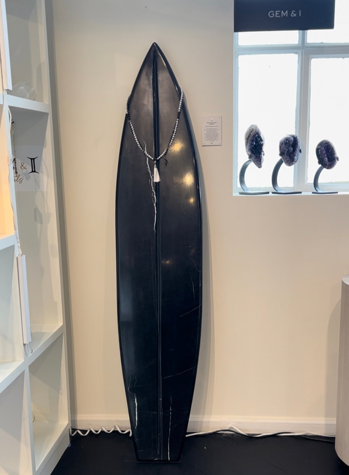 black onyx surfboard propped against beige wall near window with mala beads hanging on it