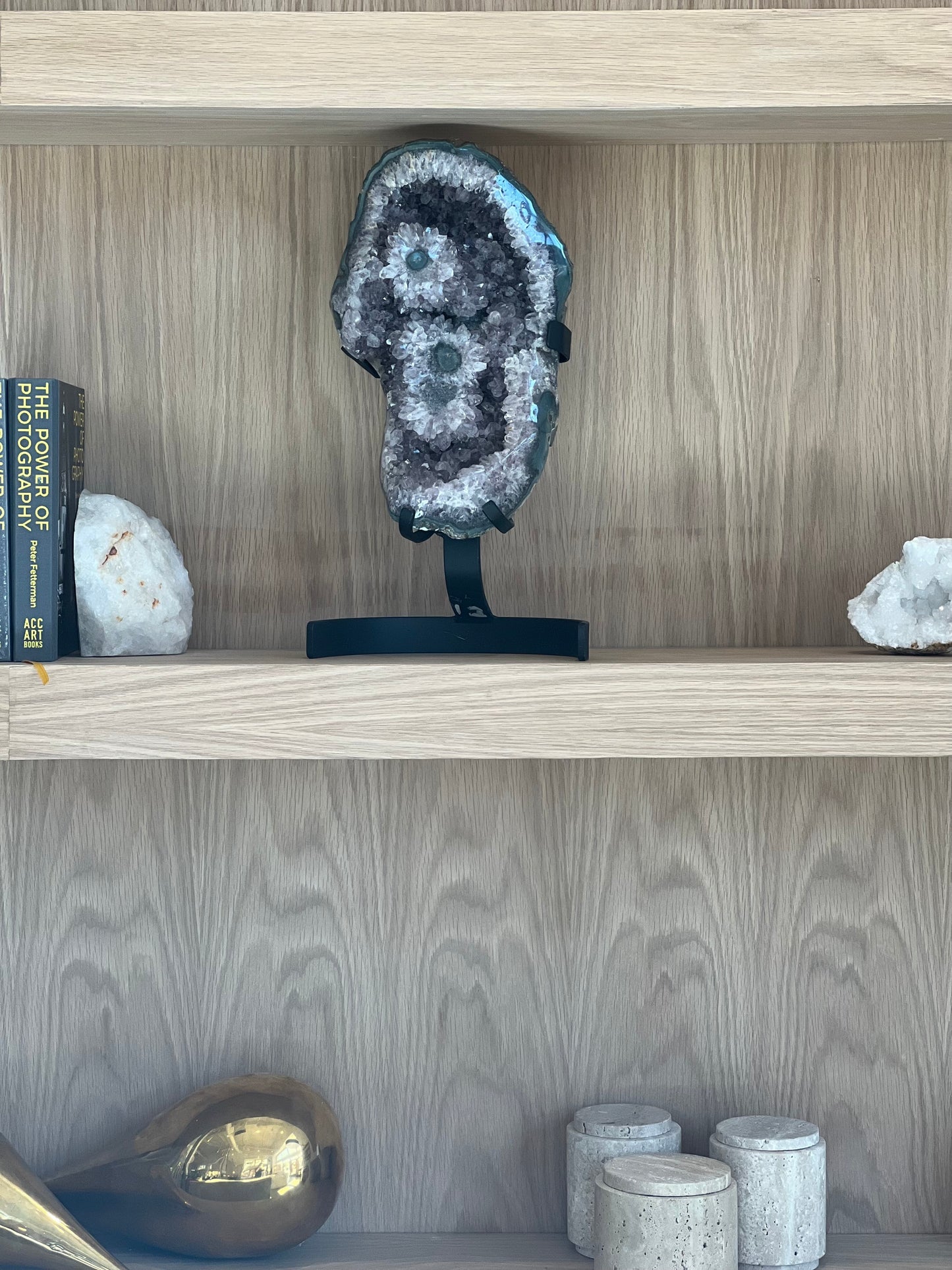 Brazilian amethyst geode featuring rare stalactite flower formations with green centers. Displayed upright on a black metal stand on wood bookshelf.