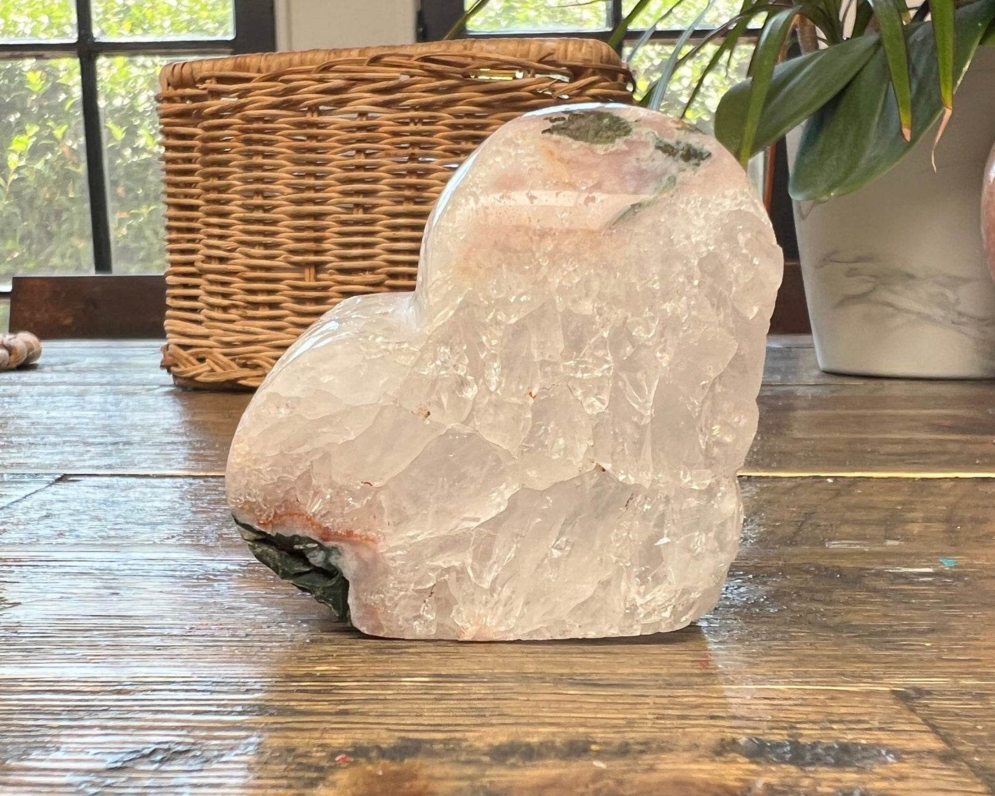 Pink Amethyst Heart shaped crystal on a wooden surface with a wicker basket and plant in the background