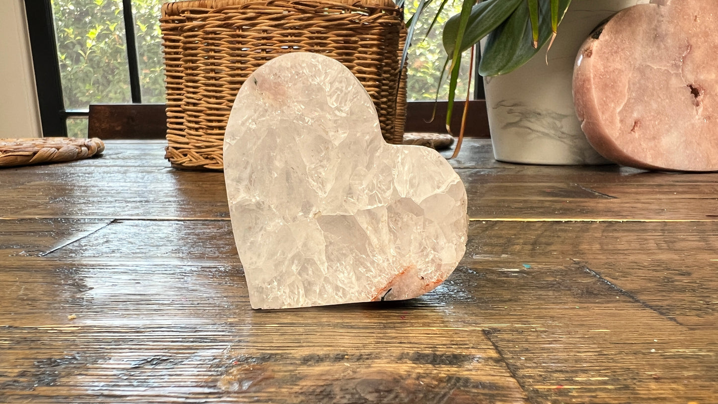 Pink Amethyst Heart shaped crystal on a wooden surface with a wicker basket and plant in the background