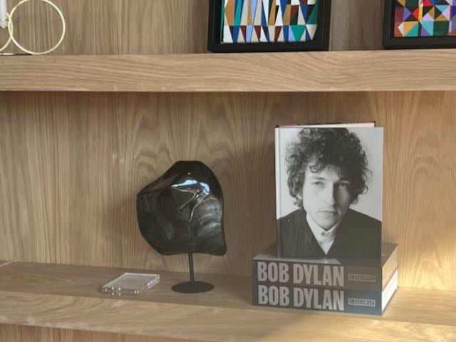 Bookshelf with a book featuring Bob Dylan on a wooden shelf. next to a large polished obsidian stone on a stand, with a silver sheen appearance.