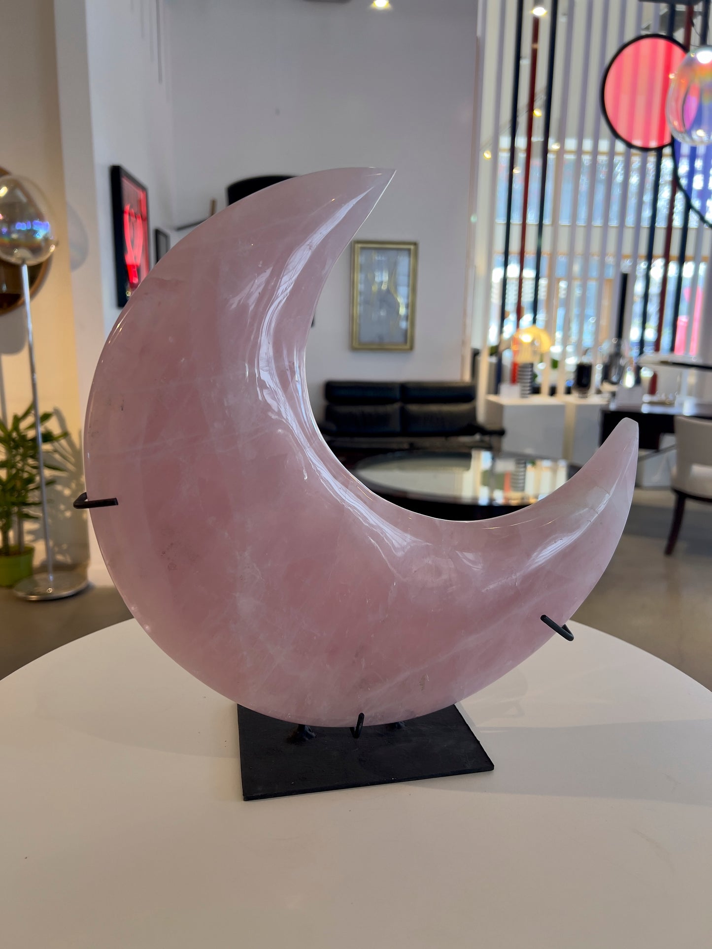 A large pink rose quartz sculpture shaped like a crescent moon, displayed on a black stand with a blurred background featuring a modern interior.