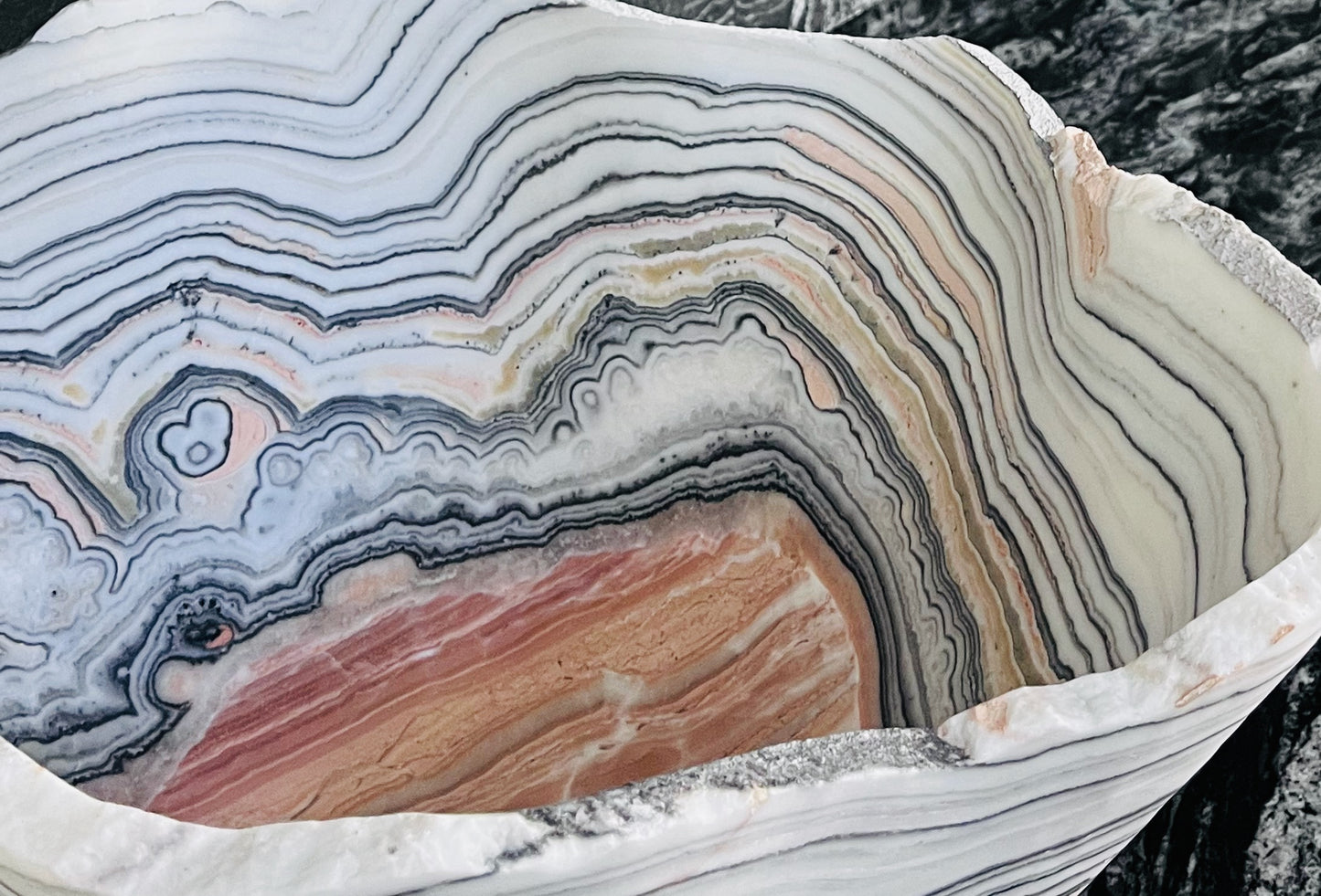 A close-up of a carved stone bowl with intricate concentric banding in cream, gray, black, and peach tones, set against a dark textured stone background.