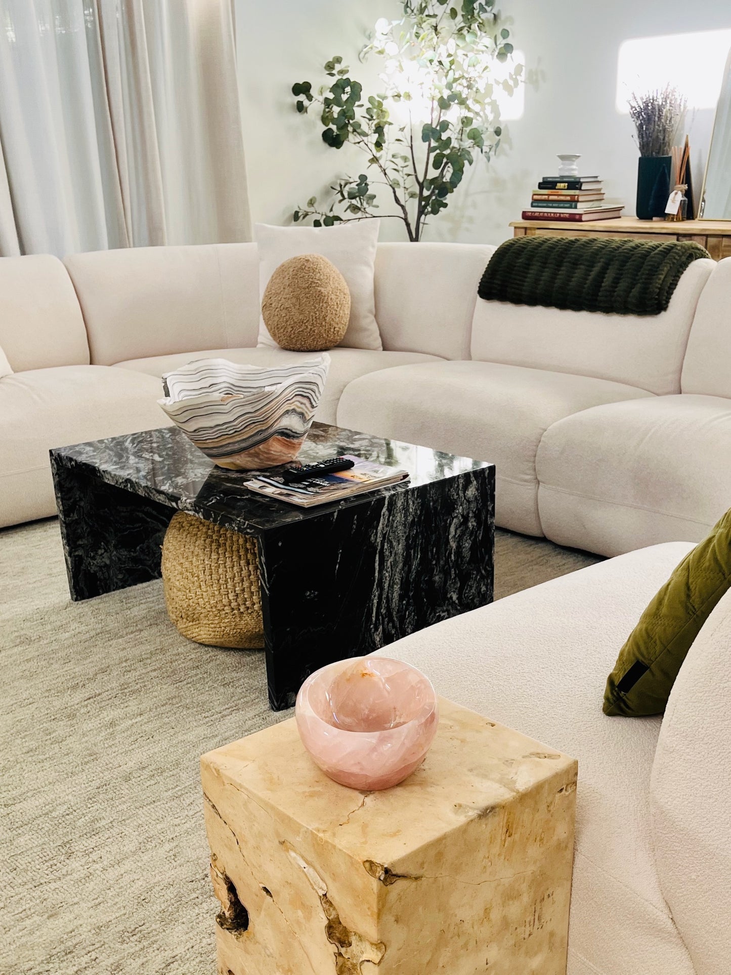 A modern living room scene featuring a cream sectional sofa, a black marble coffee table, a woven side stool, and a light wood end table in the foreground with a pink bowl.