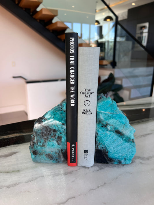 Turquoise bookends holding 2 books on a marble surface with a blurred staircase behind them.