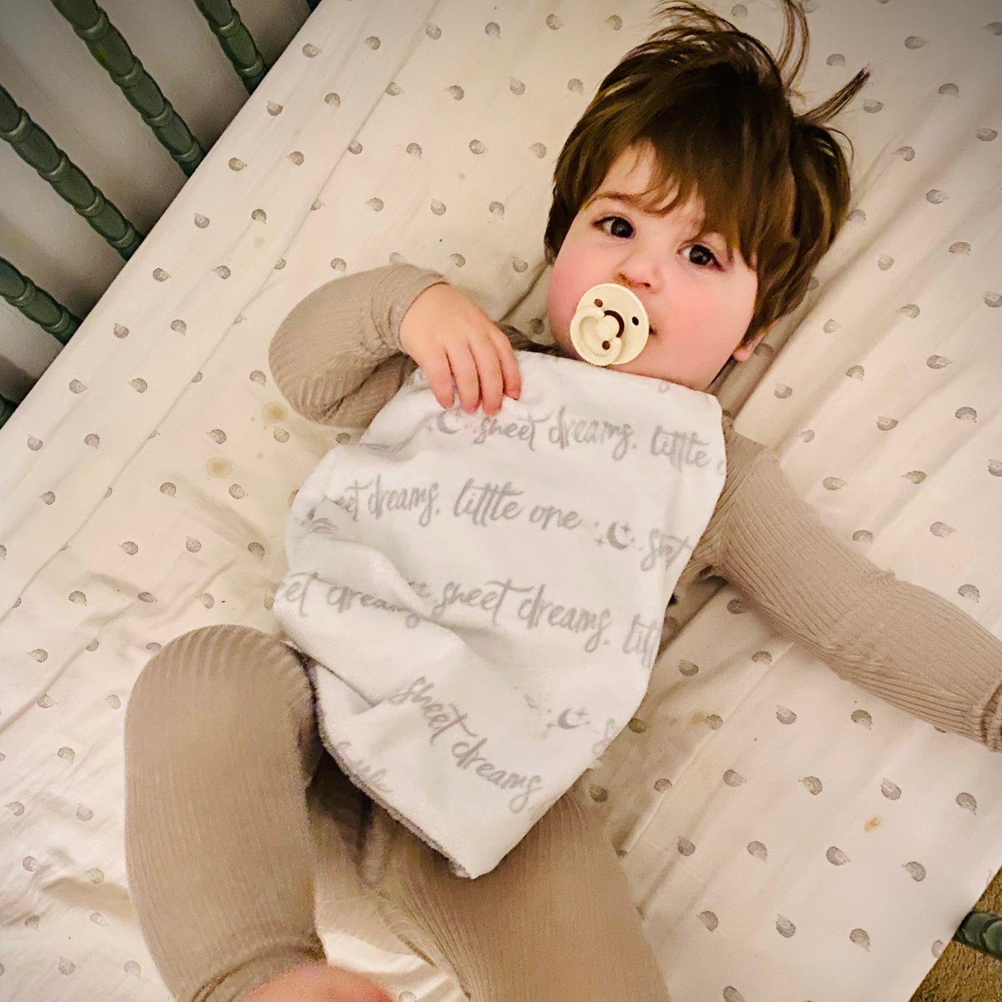 Child in a crib holding a bib and pacifier, with text on the bib.