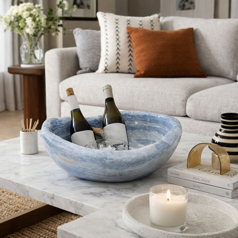 Wine bottles in a blue calcite bowl on a marble coffee table with a candle and books in a living room.