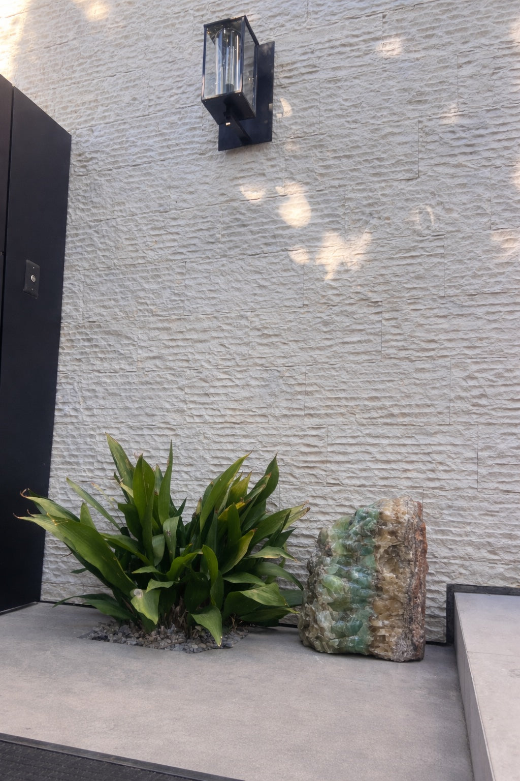 Modern outdoor setting with a textured wall, plant, and large decorative stone.