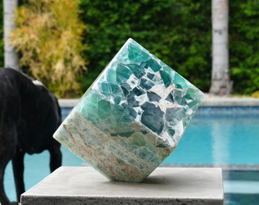 A polished Fluorite cube with a natural geometric pattern, placed on a flat surface near a pool, with a black dog in the background.
